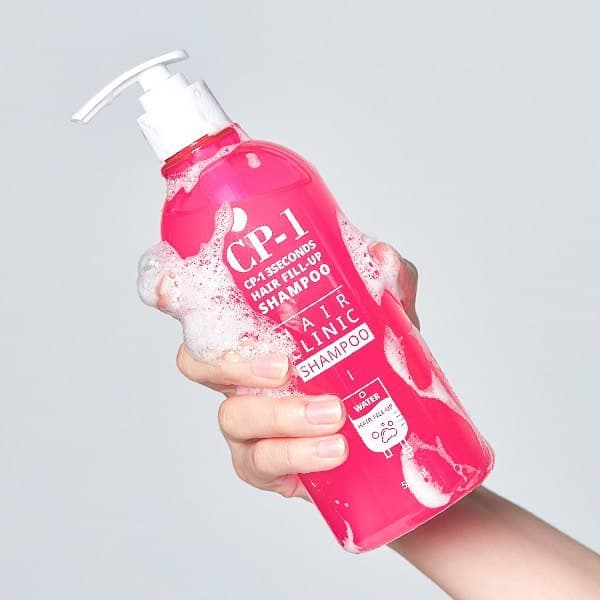 Hand holding a pink bottle of CP-1 shampoo against a gray background